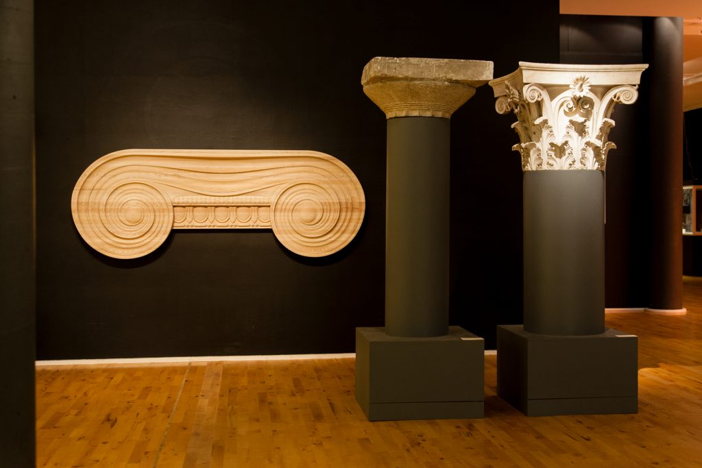Two ancient column capitals on plinths next to a wall-mounted capital-shaped artwork