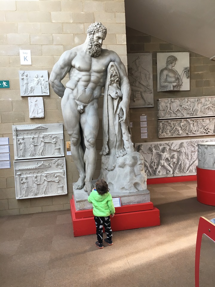 a little boy in a bright green coat stands in front of a huge sculpture of a naked bearded man