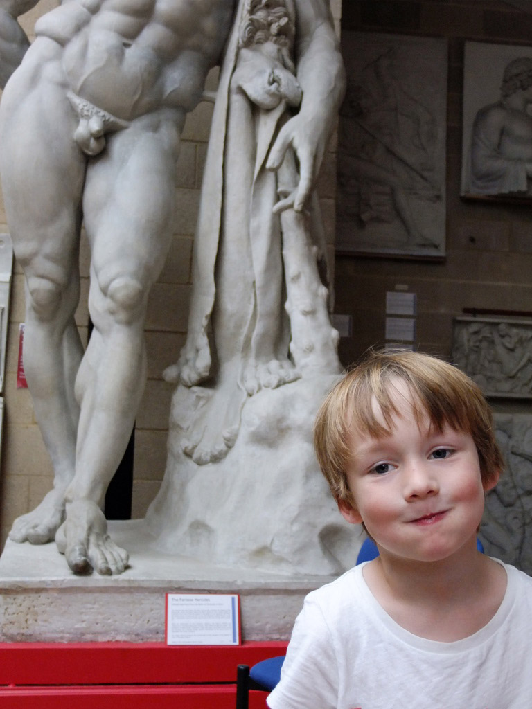 a little boy stands in front of the legs of a huge statue of a naked man