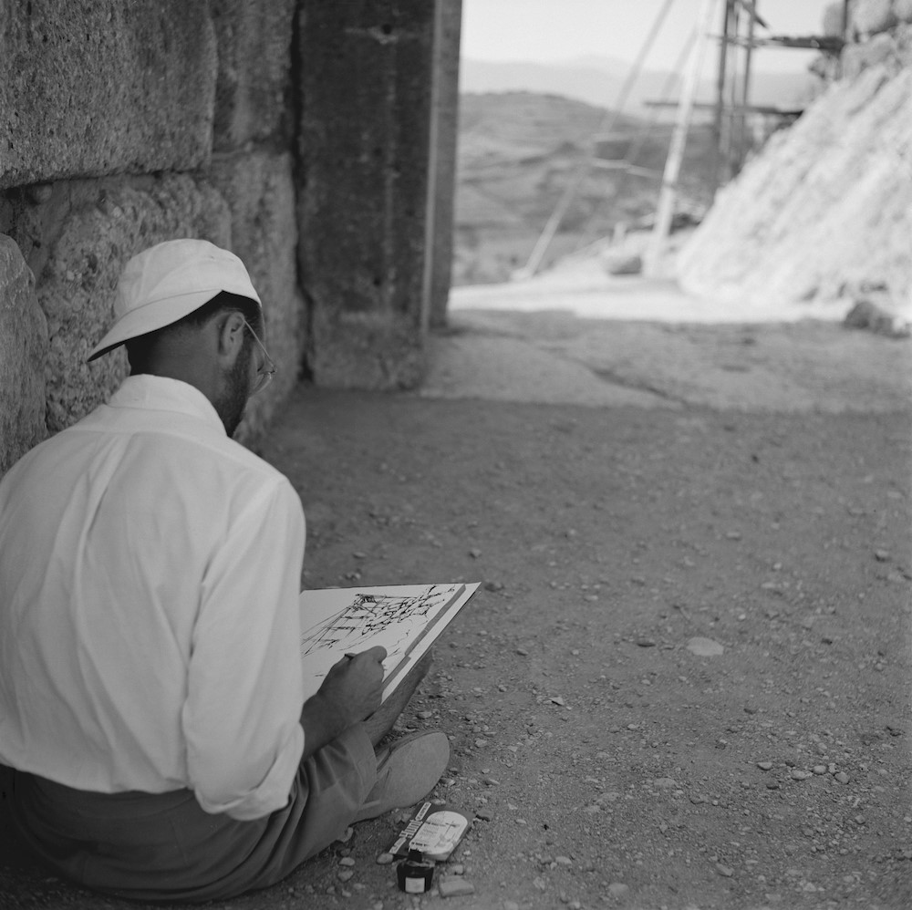 black and white image of man with back to camera sketching archeological remains