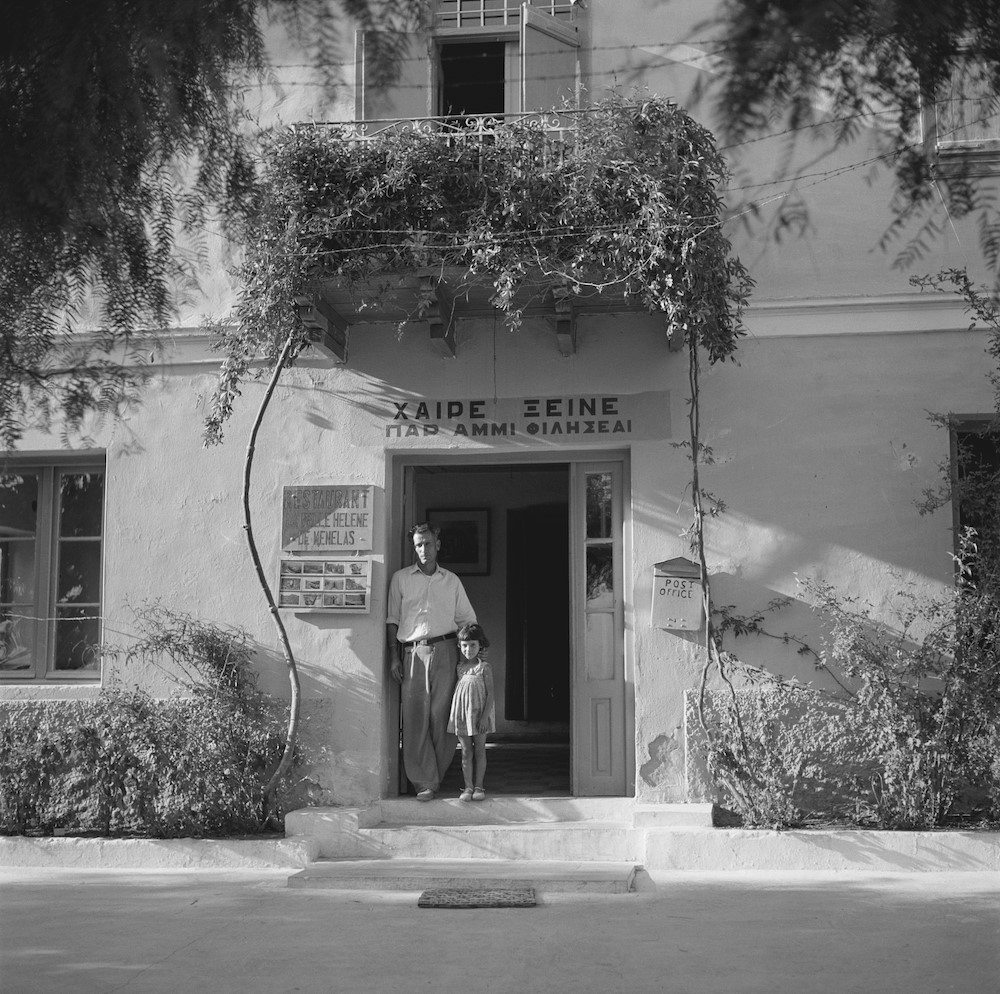 Man and young girl stand in doorway with Greek signs and flowers, black and white photo