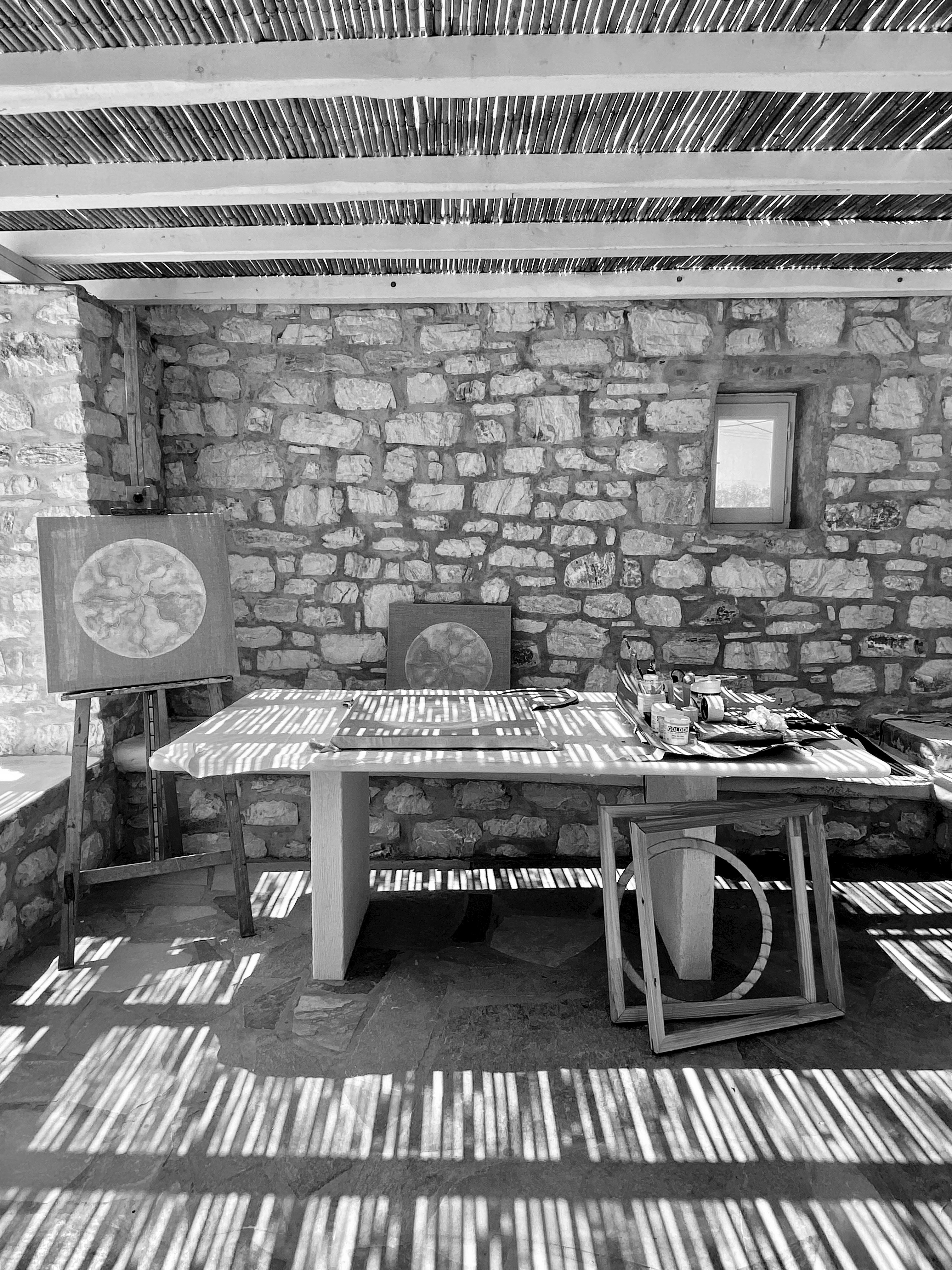 black and white view of table with circular artworks, illuminated by sunlight shining through a slatted roof-cover