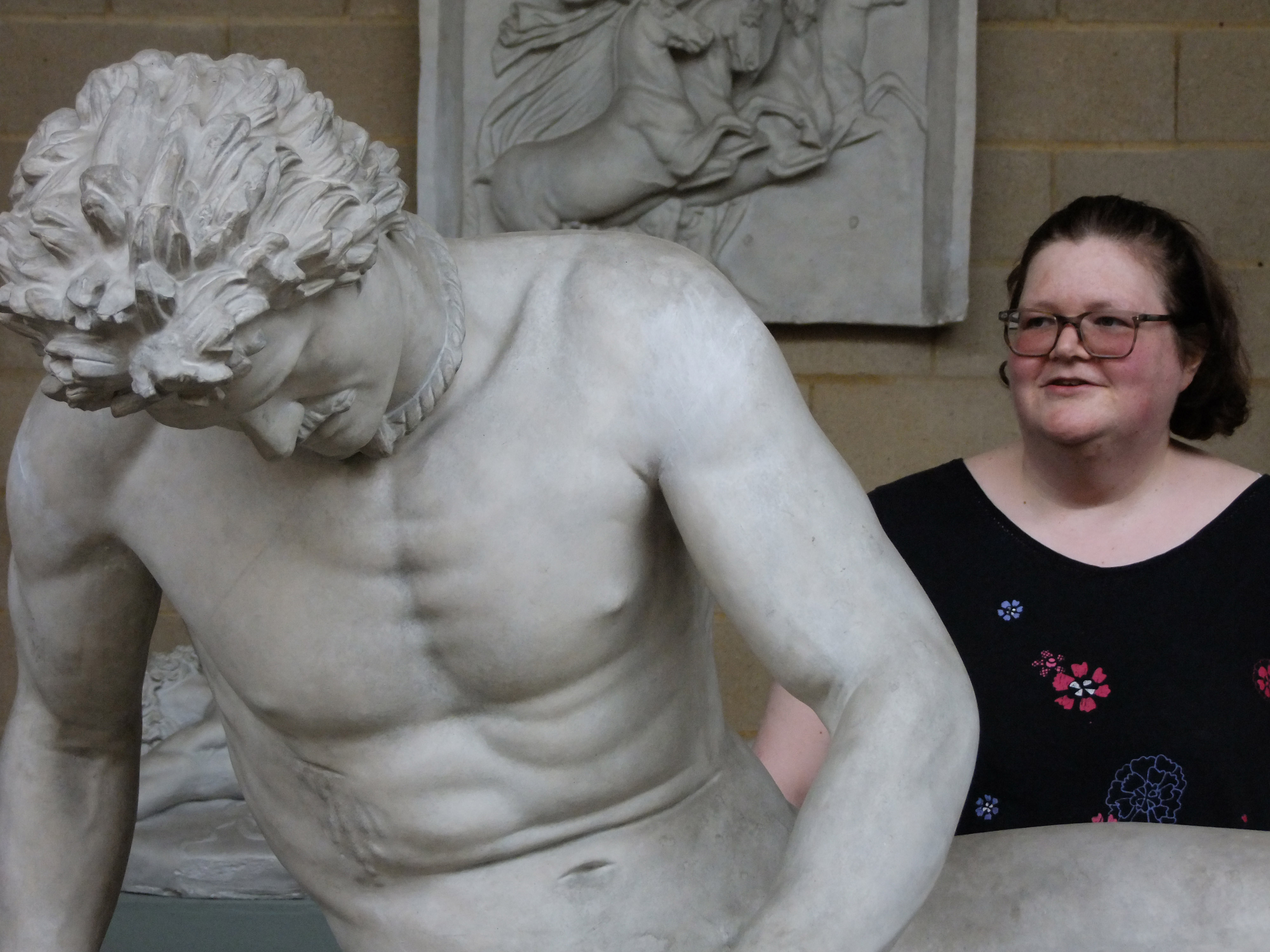 a woman in glasses stands behind a sculpted dying man with moustache