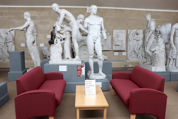 two red sofas with arm-rests next to a low table, amongst statues