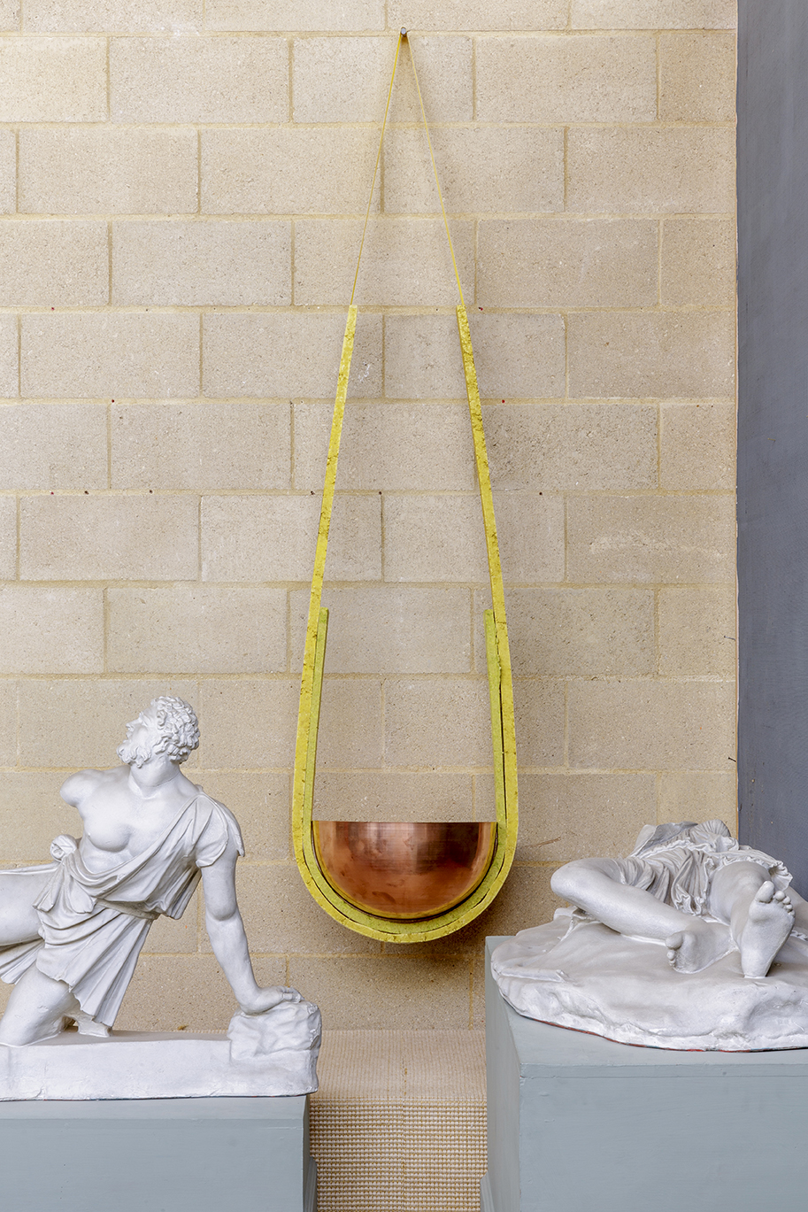 copper bowl hanging from yellow tape against breeze block wall behind two reclining plaster casts