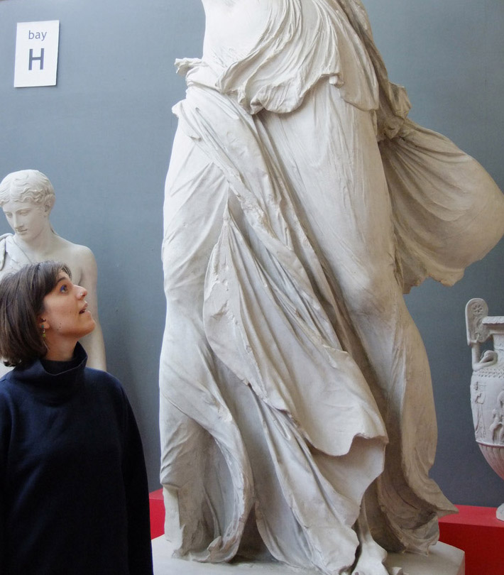 a young woman with short dark hair looks up toward a sculpted female figure