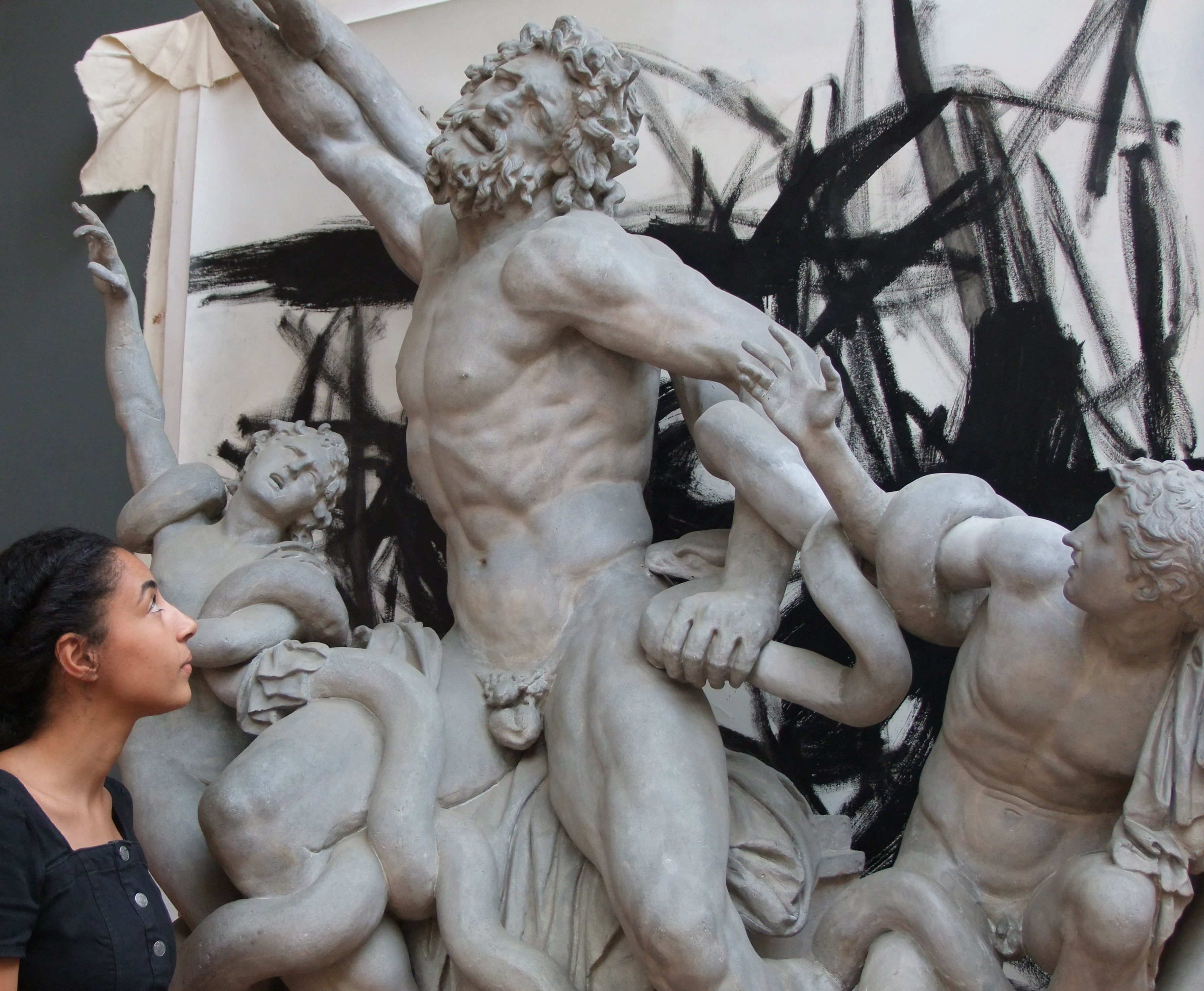 a young woman of mixed heritage looks at a sculpture of a father and two sons fighting snakes