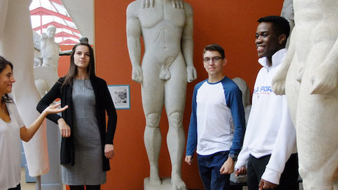 four students of different ages stand around archaic statues against an orange wall