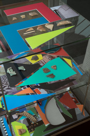 Colourful multi-media installations in glass display cabinet, with broken pottery layered on coloured paper shapes