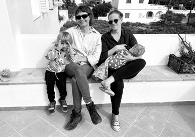 Two women sit on a white bench, one with an older child and the second nursing a baby