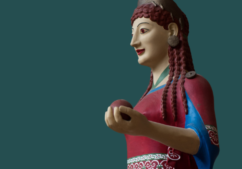 painted peplos kore, a female figure in a read dress and bronze hair ornament, holding out a round object in her hand
